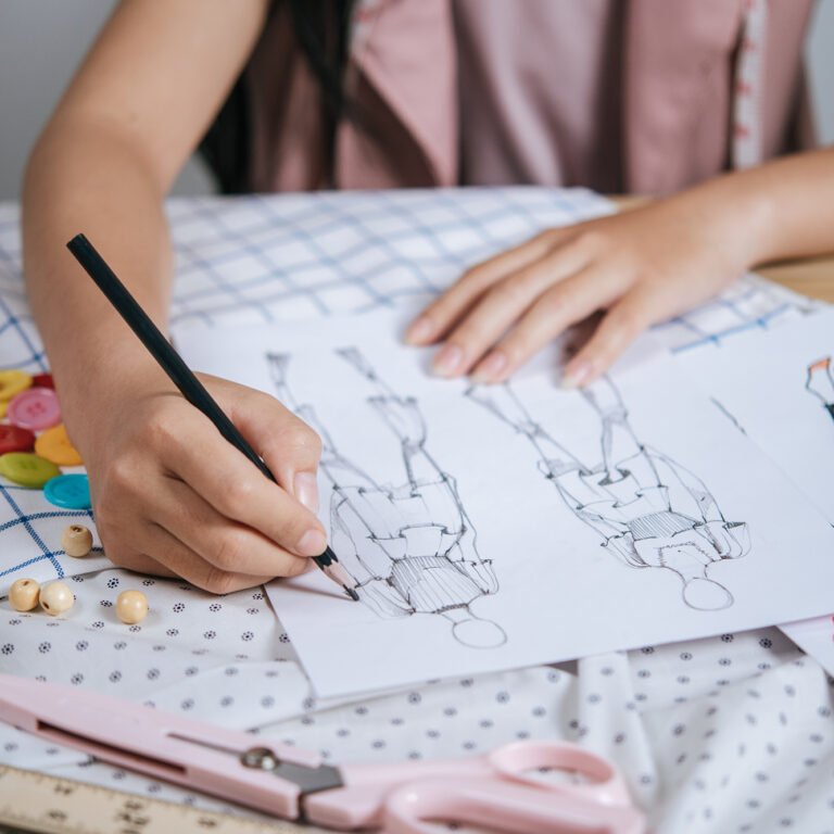 Selective focus, Close up hands of young designer or tailor sketching fashion clothes on paper, full of sewing tools on table